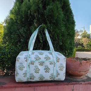 May include: A white quilted travel bag with a floral pattern in blue and brown. The bag has a rectangular shape with a top zipper and two handles. The bag is trimmed with a light blue piping.