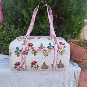 May include: A white quilted tote bag with a floral pattern in red, blue, and green. The bag has pink trim and handles. The bag is rectangular with a pocket on the front.