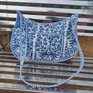 Puede incluir: Bolso de hombro con estampado floral azul y blanco. El bolso presenta un diseño floral repetido en tonos azules sobre un fondo blanco. Tiene un borde a rayas y una correa a juego.