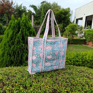 May include: A large tote bag with a floral pattern in shades of blue, pink, and green. The bag has pink handles and trim, and the floral design features lotus flowers. The bag is sitting on a green bush.