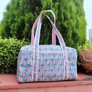 May include: A blue quilted tote bag with a floral pattern in pink and white. The bag has pink and white striped handles and trim. The bag has a rectangular shape with a pocket on the front.