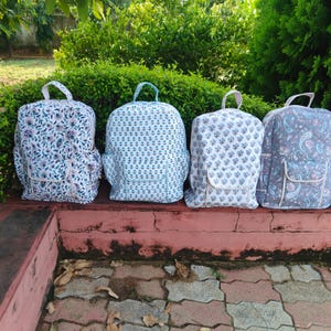 Könnte beinhalten: Vier gesteppte Stoffrucksäcke in einer Reihe. Jeder Rucksack hat einen Tragegriff, eine Vordertasche mit Klappe und ein florales oder Paisley-Muster in Blau-, Weiß- und zarten Rosatönen. Die gesteppte Textur ist auf allen Taschen deutlich erkennbar.