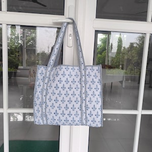 May include: A white and blue patterned tote bag with matching straps hangs on a white door. The bag features a repeating floral design in blue, with a striped border. The bag is made of fabric and appears to be handmade.
