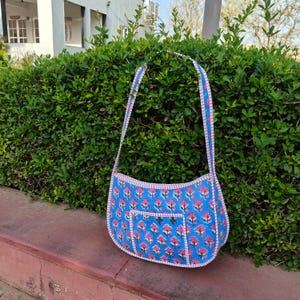 May include: A blue shoulder bag with a floral pattern in pink and white. The bag has a small front pocket and a matching strap. The bag is sitting on a red surface in front of a green hedge.