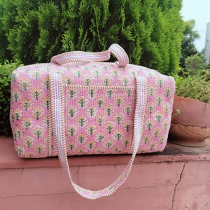 Puede incluir: Una bolsa de viaje acolchada rosa con un estampado floral en amarillo y verde. La bolsa tiene forma rectangular con un asa superior y una correa larga para el hombro. Los bordes están ribeteados con tela a rayas blancas y doradas.