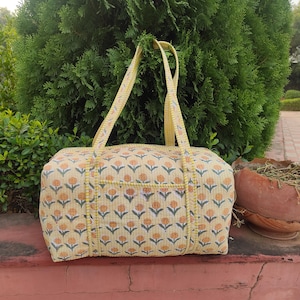 May include: A yellow floral print tote bag with matching yellow handles. The bag has a repeating pattern of orange flowers and green leaves. The bag is on a red surface in front of a green bush.