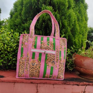 May include: A pink and green tote bag with a tiger print design. The bag features vertical green and pink stripes, a zipper pocket, and pink handles. The tiger print is in shades of brown and tan.