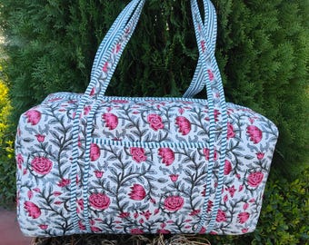 Pode incluir: Uma sacola tote com estampa floral com rosas cor-de-rosa e folhas verdes em um fundo branco. A sacola tem alças e acabamentos listrados em azul e branco. A sacola é feita de tecido acolchoado.