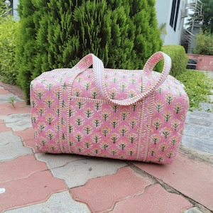 May include: Pink floral patterned duffel bag with white and beige trim. The bag has two handles and a zipper closure. The bag is sitting on a red and pink tiled surface. The bag is made of a soft, quilted fabric.