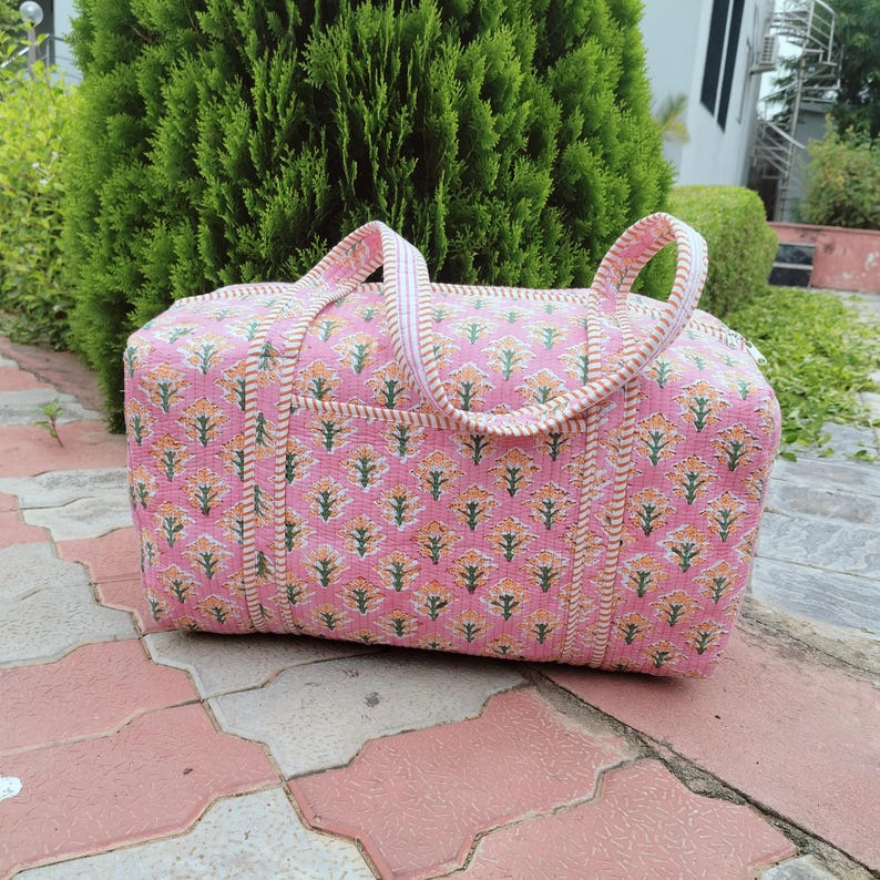 May include: Pink floral patterned duffel bag with white and beige trim. The bag has two handles and a zipper closure. The bag is sitting on a red and pink tiled surface. The bag is made of a soft, quilted fabric.