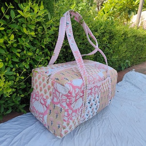 Peut inclure: Un sac de voyage matelassé rectangulaire présentant un motif patchwork avec divers imprimés floraux et botaniques dans des tons de rose, blanc et beige. Le sac comprend deux longues anses en tissu à rayures roses et blanches et une fermeture éclair.