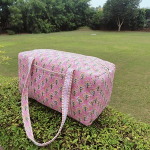 May include: A pink floral print travel bag with a rectangular shape. The bag has two handles and a pocket on the front. The floral pattern includes yellow and green accents. The bag is sitting on a green bush.