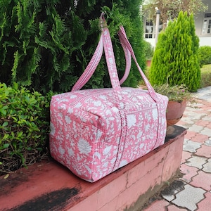 Puede incluir: Una bolsa de lona rosa y blanca con estampado floral y correas a juego. La bolsa es rectangular con apariencia acolchada. Las correas son lo suficientemente largas para llevar al hombro. La bolsa está sobre una superficie roja.