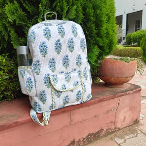 Op de afbeelding: Een gewatteerde stoffen rugzak met een witte basis en een herhalend blauw bloemenpatroon. De tas heeft een voorvak met klep, zijvakken met groene bies, een handvat aan de bovenkant en verstelbare schouderbanden met metalen gespen.