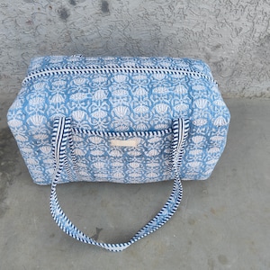 Puede incluir: Una bolsa de lona con estampado floral azul y blanco con asas a rayas. La bolsa tiene forma rectangular con cierre de cremallera y un pequeño bolsillo en la parte delantera. La bolsa está hecha de tela acolchada.
