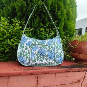 Puede incluir: Bolso de hombro con estampado floral blanco y azul, con ribete y correa verdes. El bolso tiene un bolsillo frontal grande y está colocado sobre un fondo de follaje verde y una superficie roja. El diseño del bolso presenta flores azules.