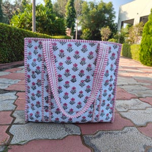 Może przedstawiać: Biała torba typu tote z różowym kwiatowym wzorem i różowymi paskami. Torba ma dwa uchwyty i stoi na ceglanej powierzchni. Kwiatowy wzór powtarza się na całej torbie.