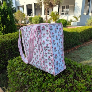 May include: A white tote bag with a floral pattern in pink and green. The bag has pink trim and handles. The bag is sitting on a green bush.