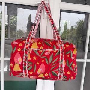 May include: A red tote bag with a floral and bird pattern. The bag features a rectangular shape with a front pocket and two handles. The pattern includes yellow flowers, green leaves, and pink and yellow birds. The handles are striped with red and white.