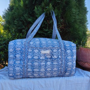 May include: A rectangular quilted duffel bag featuring a blue and white floral block print pattern. The bag includes two long shoulder straps with a navy and white striped design, a front slip pocket, and a small rectangular label on the front.