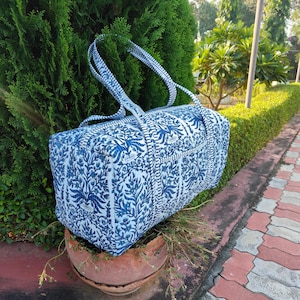 May include: A blue and white patterned duffel bag with matching handles. The bag features a floral design and is sitting on a terracotta pot. The bag is rectangular and appears to be made of fabric.