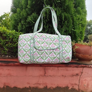 Può includere: Borsa tote rettangolare con un motivo floreale rosa e verde su sfondo bianco. La borsa ha manici e finiture verdi. La borsa è appoggiata su una superficie rossa con fogliame verde sullo sfondo.