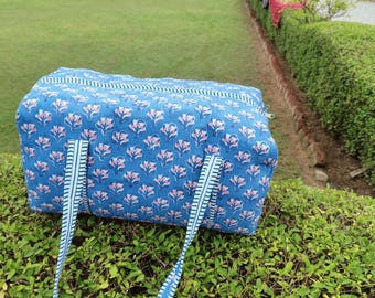 May include: A blue floral patterned travel bag with white and blue striped handles. The bag is rectangular with a zipper closure and is sitting on a green bush. The floral pattern includes pink and white flowers.