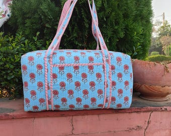 May include: A light blue quilted tote bag with a floral pattern of red flowers. The bag has red and white striped handles and trim. The bag is rectangular with a small pocket on the front.