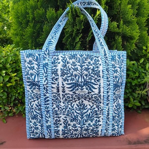 May include: A blue and white tote bag with a floral pattern. The bag has a rectangular shape with a matching blue and white striped trim and handles. The bag is made of a quilted fabric.