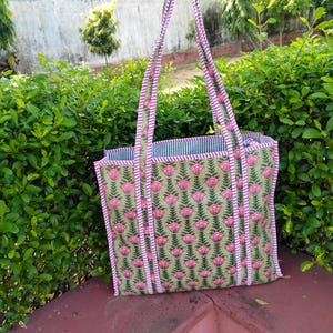 Puede incluir: Un bolso tote verde con un estampado floral rosa y ribetes a rayas rosas. El bolso tiene dos asas y un interior a rayas. El bolso está sobre una superficie roja frente a follaje verde.