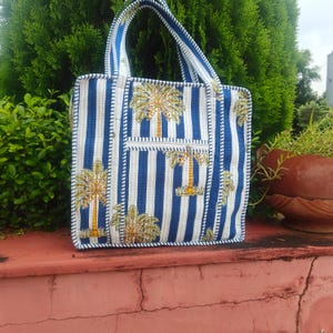 May include: A blue and white striped tote bag with a palm tree design. The bag has a rectangular shape with a small front pocket and white stitching around the edges. The handles are also striped.