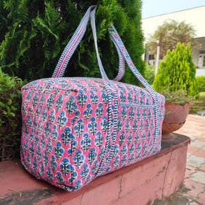 May include: A pink and blue floral patterned travel bag with matching straps. The bag has a rectangular shape with a zipper closure and is made of quilted fabric. The straps are white with pink and blue stripes.