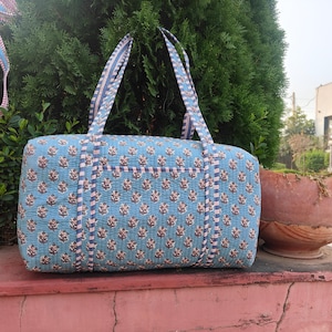 Puede incluir: Un bolso tote acolchado azul con un estampado floral en marrón y blanco. El bolso tiene dos asas y un bolsillo en la parte delantera. El bolso mide aproximadamente 46 cm de largo.