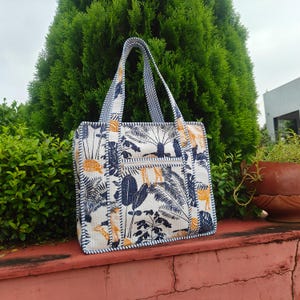 Könnte beinhalten: Eine weiße Tragetasche mit tropischem Aufdruck mit Palmen, Giraffen und anderem Laub in Marineblau und Gelb. Die Tasche hat eine Reißverschlusstasche und blau-weiß gestreifte Griffe. Die Tasche ist etwa 38 cm breit und 30 cm hoch.