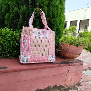 May include: A rectangular tote bag featuring a patchwork design with various floral and botanical block-print patterns in shades of pink, white, and tan. The bag has two matching fabric handles and a quilted texture throughout the exterior surface.