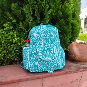 May include: A teal and white patterned backpack featuring a repeating botanical print. The bag includes a front pocket with a rounded flap, a side pocket containing a clear plastic water bottle, and adjustable shoulder straps made of the same fabric.