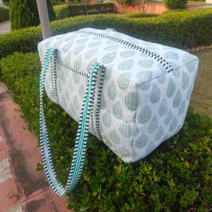 May include: A white and green leaf-patterned tote bag with striped handles. The bag is rectangular with a zipper closure and is made of quilted fabric. The handles are a combination of black, white, and green stripes.