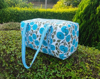 Pode incluir: Uma mala de viagem retangular com fundo branco e um padrão repetido de flores turquesas e folhas verdes. A mala tem alças e acabamentos turquesa. A mala está colocada em um arbusto verde.