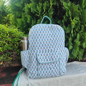 Könnte beinhalten: Ein gesteppter Stoffrucksack mit einem sich wiederholenden Muster aus kleinen blauen Blumen auf weißem Grund. Der Rucksack verfügt über eine Vordertasche mit Klappe, einen Tragegriff und Seitentaschen, von denen eine eine silberne Metallflasche enthält.