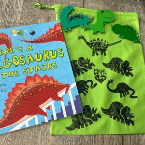 May include: A children's book titled "There's a Stegosaurus on the Stairs" with a red and blue cover, next to a green drawstring bag decorated with black dinosaur silhouettes and wooden dinosaur cutouts. The book's cover features a red stegosaurus.