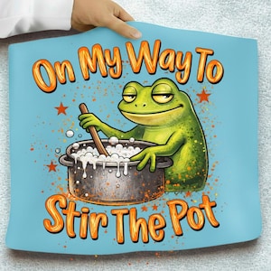 May include: A light blue decorative pillow featuring a cartoon frog stirring a pot. The text "On My Way To Stir The Pot" is written in orange. The frog is green and the pot is dark grey.