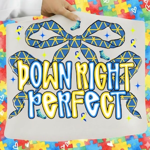 Peut inclure: Un sac fourre-tout blanc avec l'inscription "DOWN RIGHT PERFECT" en jaune et bleu, entourée d'un motif de nœud bleu. Le motif comprend des chaussettes bleues et des étoiles. Le fond présente un motif de puzzle coloré.
