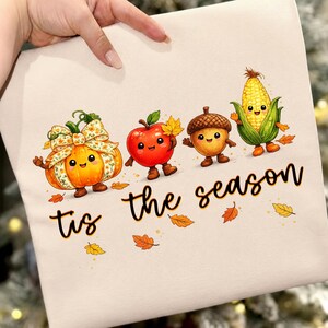 Peut inclure: Un t-shirt de couleur claire avec un motif représentant une citrouille souriante avec un nœud floral, une pomme rouge, un gland et un épi de maïs avec des visages. Le texte 'tis the season' est imprimé en écriture cursive noire sous les personnages.