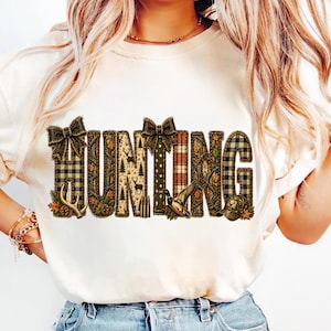 Peut inclure: Un t-shirt couleur crème avec le mot HUNTING en grandes lettres majuscules. Chaque lettre est remplie de motifs incluant du tartan, du camouflage, des cerfs, des arbres, des balles, un canard et une casquette, ornés de deux nœuds marron.