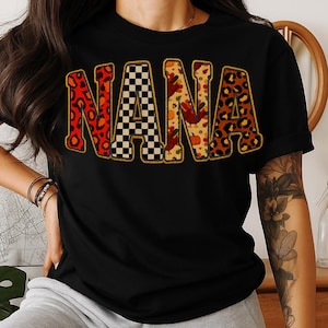Peut inclure: T-shirt noir avec le mot "NANA" en grandes lettres colorées. Les lettres présentent différents motifs : imprimé léopard, damier noir et blanc, et un motif sur le thème de l'automne avec des citrouilles et des dindes.