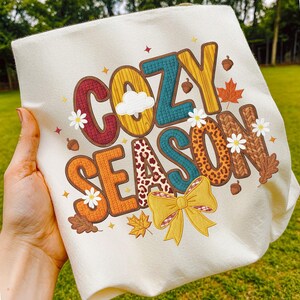 Peut inclure: Tissu crème avec l'inscription "COZY SEASON" en lettres colorées et texturées. Le motif comprend un nuage, des marguerites, des glands, des feuilles d'érable et un nœud jaune. Les lettres ont un aspect bois, léopard et tricot.