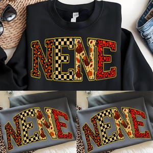 Peut inclure: Sweat-shirt noir avec le mot "NENE" dans une police décorative audacieuse. Les lettres présentent un mélange d'imprimé léopard, de motif damier noir et blanc et d'illustrations sur le thème de l'Action de grâce avec des dindes et des feuilles d'automne. Le sweat-shirt est présenté sur une surface blanche.