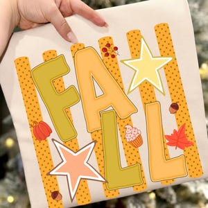 Peut inclure: Un t-shirt couleur crème arborant le mot FALL en grandes lettres bloc jaunes et orange. Le motif comprend des rayures verticales orange à pois, deux étoiles, une citrouille rouge, un petit cupcake, une branche de baies et deux glands marron.