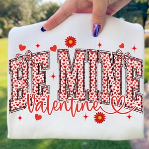 Peut inclure: Sweat-shirt blanc avec l'inscription "BE MINE Valentine" en rouge, remplie de cœurs. Le motif comprend des cœurs rouges, des nœuds et des motifs floraux, parfait pour la Saint-Valentin.