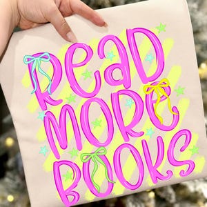 Peut inclure: Un sac fourre-tout beige avec l'inscription "READ MORE BOOKS" en rose vif, soulignée de rose foncé. Le texte est orné de nœuds et d'étoiles de différentes couleurs, sur un fond jaune.